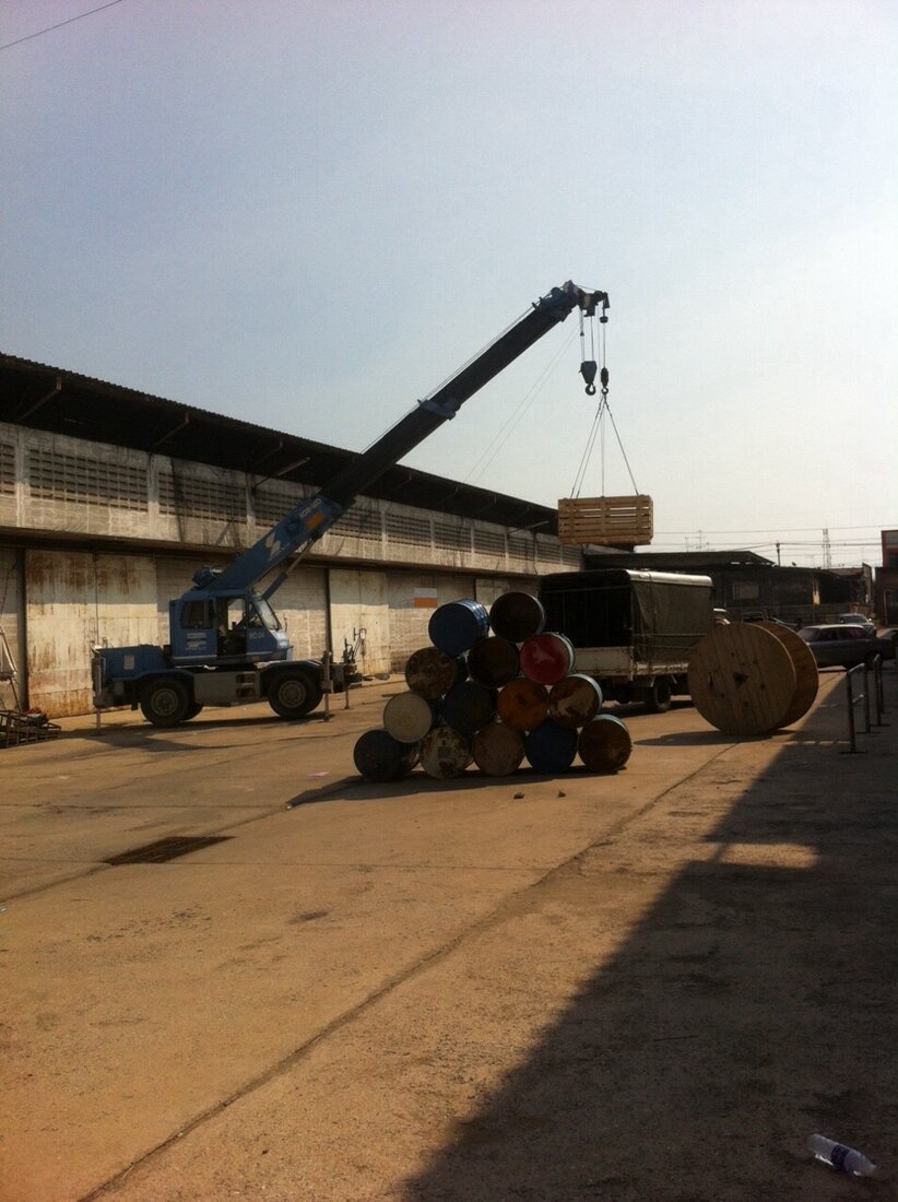 เครนขนาดเล็กของ BIG MACHINE TRADING ทำงานที่กองถ่ายโฆษณา ท่ามกลางถังน้ำมันและม้วนสายเคเบิลไม้ที่ใช้เป็นอุปกรณ์ประกอบฉาก