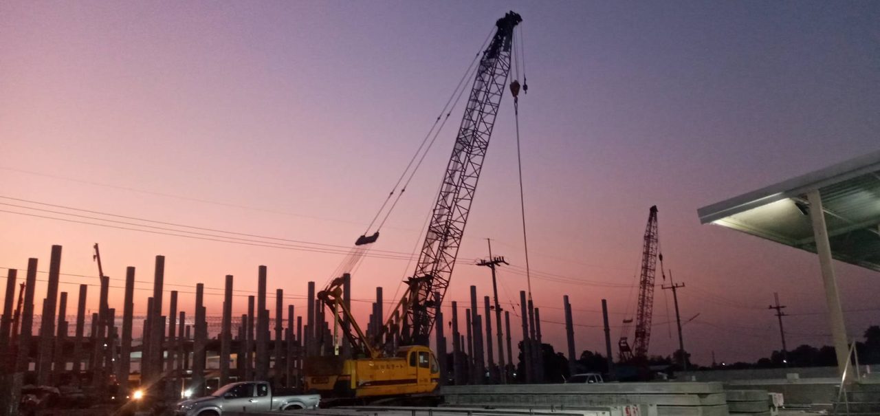 เครนตีนตะขาบสีเหลืองของ BIG MACHINE TRADING ทำงานยามพระอาทิตย์ตกที่บ้านบึง ชลบุรี ท่ามกลางเสาคอนกรีตเรียงแถวในไซต์งาน