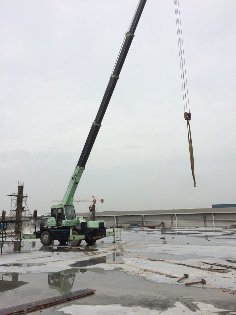 มุมใกล้ของเครนล้อยาง 10 ตัน KATO ที่ถูกเครน 25 ตันของ BIG MACHINE TRADING ยกข้ามเข้าไปในพื้นที่ก่อสร้างที่ล้อมรอบด้วยนั่งร้าน