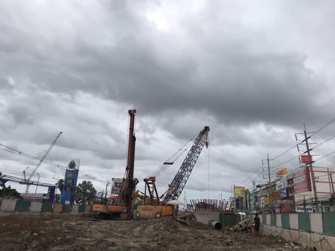 เครนตีนตะขาบ Kobelco และเครื่องเจาะเข็ม Sany ทำงานร่วมกันริมถนน ใกล้อาคารพาณิชย์ วันฟ้าใส