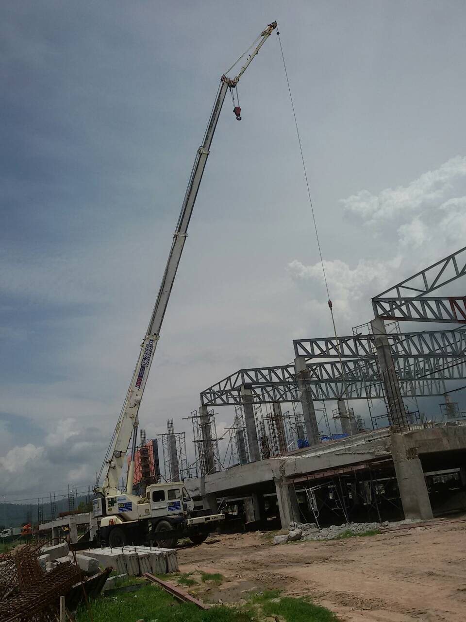 เครนล้อยางสีขาวของ BIG MACHINE TRADING ทำงานใกล้โครงเหล็กและเสาคอนกรีต ในไซต์งานก่อสร้างอาคารแสดงสินค้านักท่องเที่ยว ริมบายพาสชลบุรี