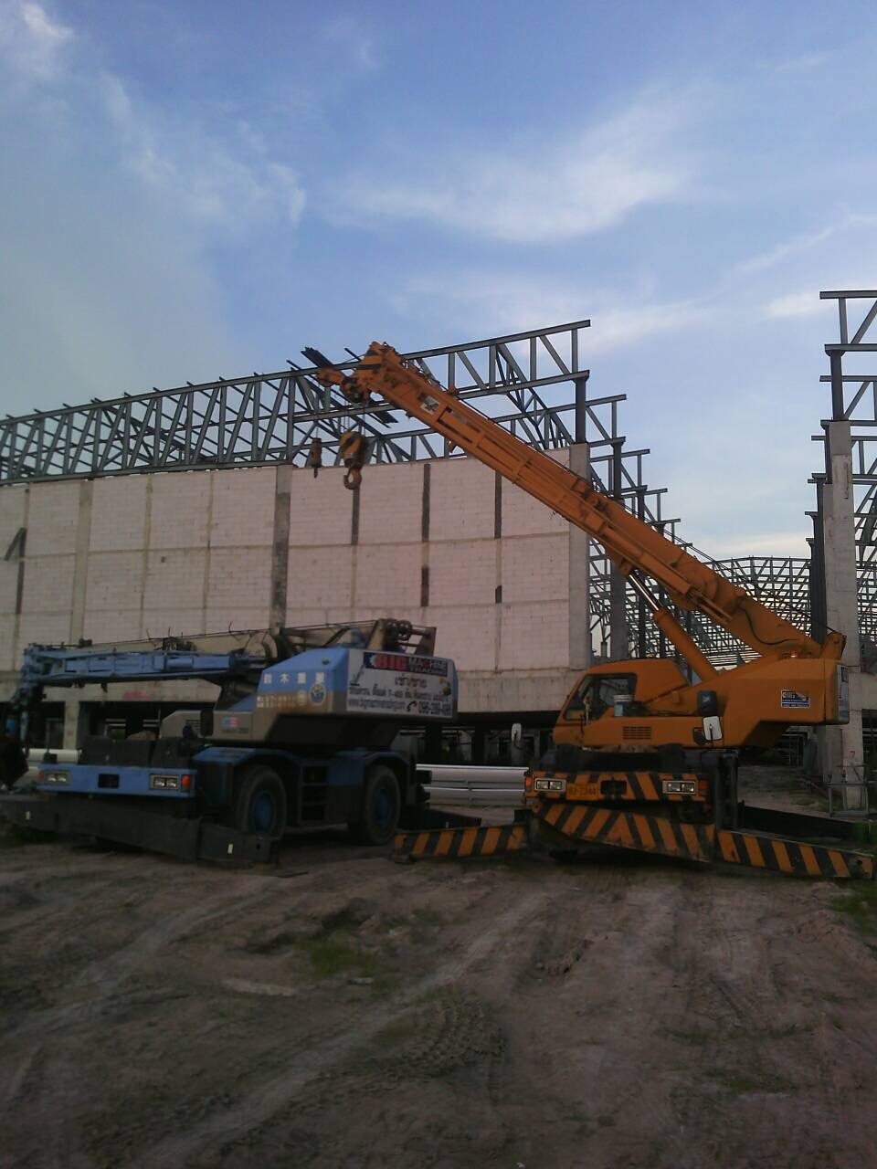 เครนล้อยางสีเหลืองของ BIG MACHINE TRADING ทำงานใกล้กำแพงบล็อกและโครงเหล็ก ในไซต์งานก่อสร้างอาคารแสดงสินค้านักท่องเที่ยว ริมบายพาสชลบุรี