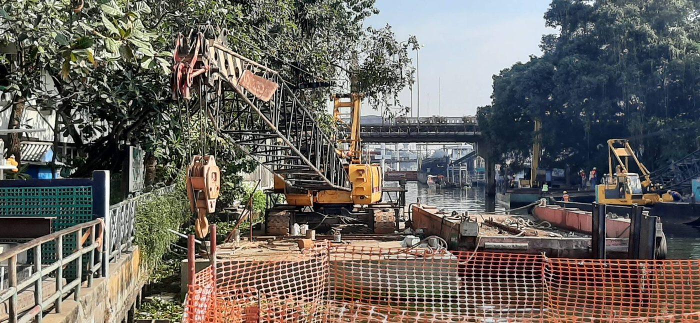 เครนโครงถักตัวเก่ายืนใต้สะพานข้ามคลอง ตะขอและลูกรอกอยู่เบื้องหน้า เห็นโป๊ะเรือและทีมงานทำงานอีกจุดหนึ่งด้านหลัง
