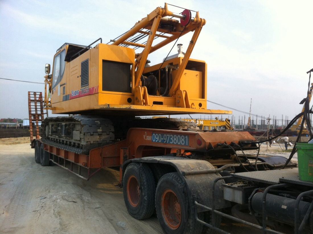 เครนตีนตะขาบ XCMG สีเหลืองบนรถเทรลเลอร์ พร้อมทางลาดสำหรับขึ้น-ลง หน้างานก่อสร้าง