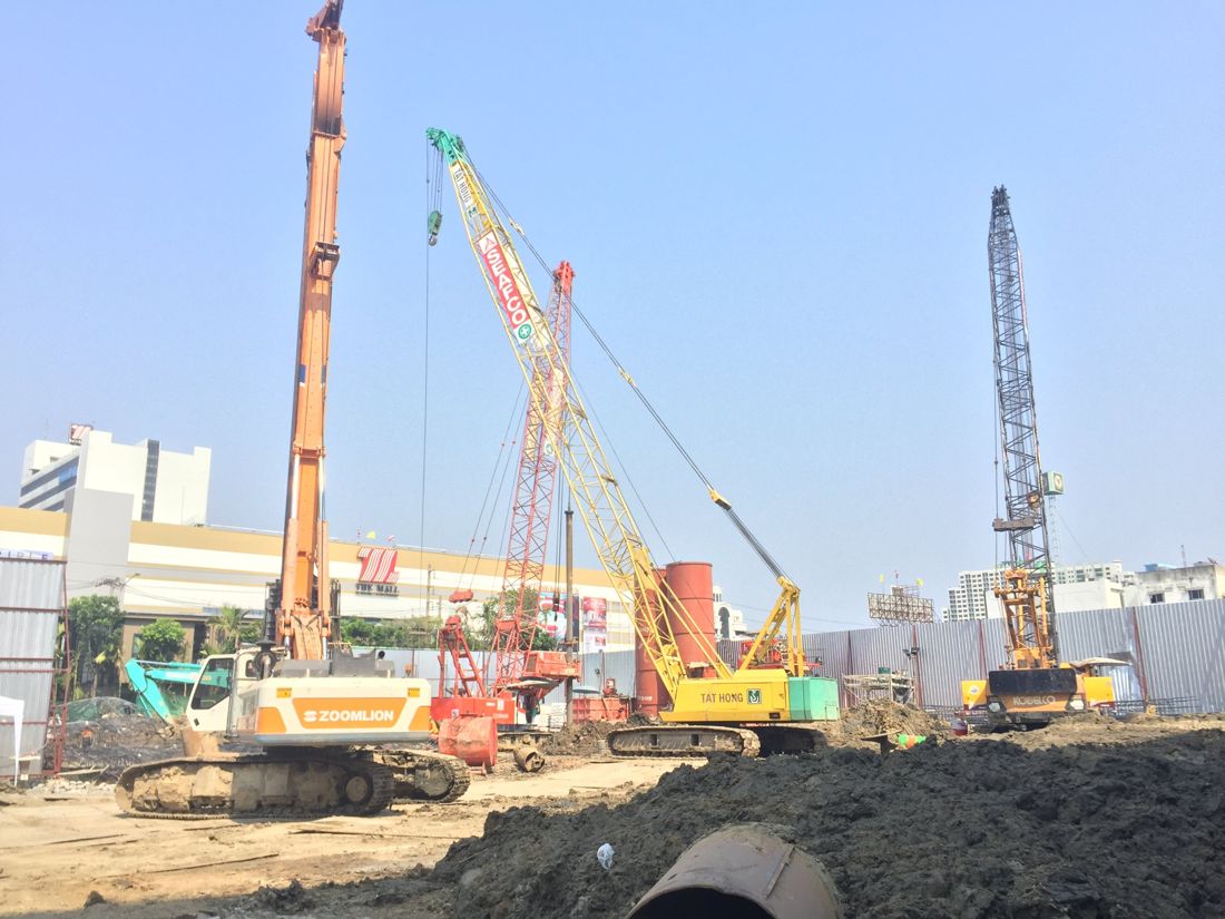 ภาพรวมไซต์งานเข็มเจาะเปียก Whizdom Station Ratchada-Thapra มีรถเจาะเข็ม เครนตีนตะขาบหลายคัน และปลอกเหล็กเสาเข็ม ด้านหลังเป็นเดอะมอลล์ ท่าพระ